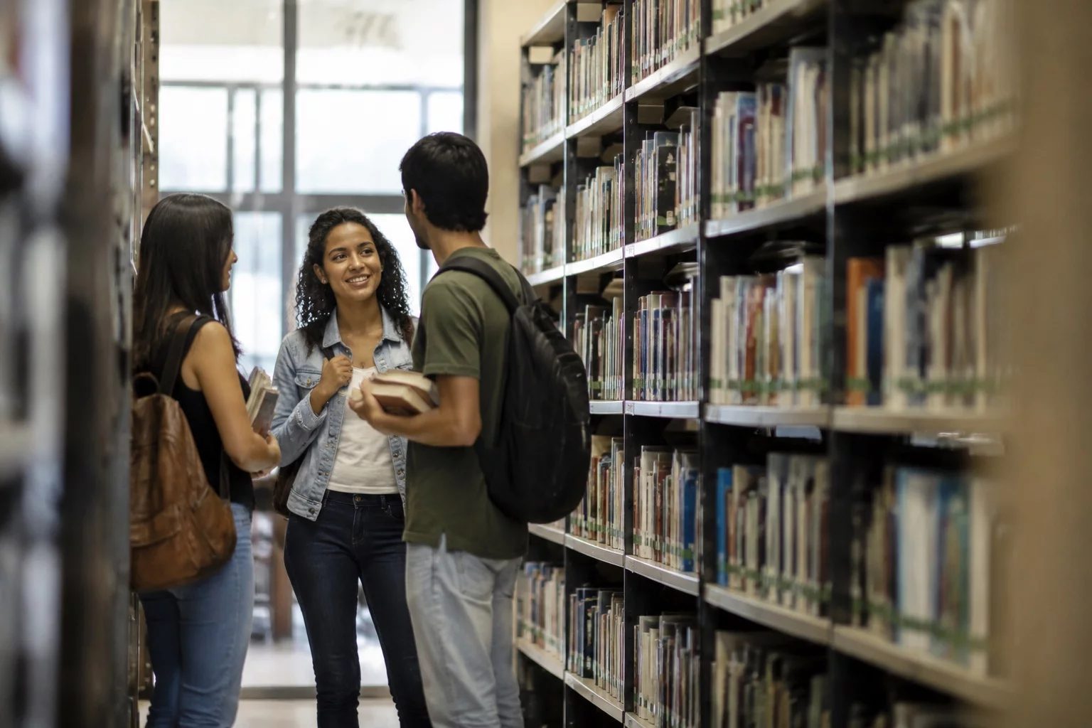 Estudantes conversando em pé entre as estantes de uma biblioteca